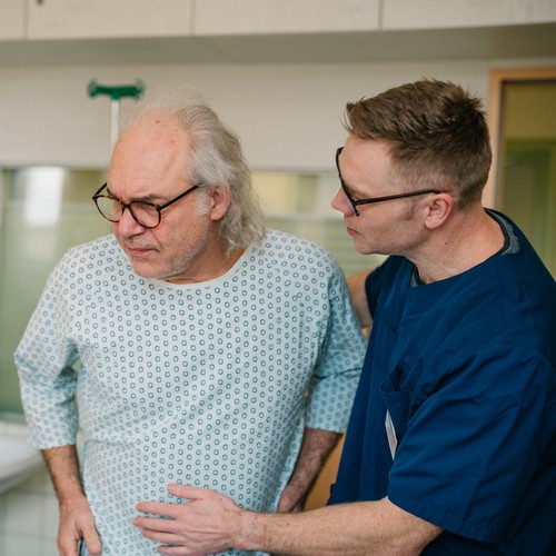 Ein junger Pfleger hilft einem älteren Patienten im Kliniknachthemd.