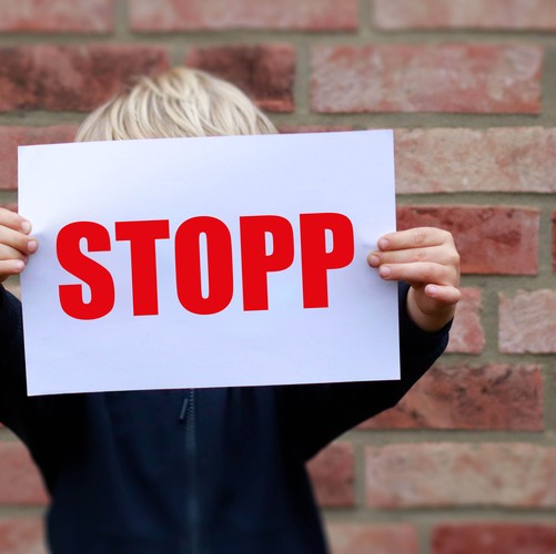Das Foto zeigt ein Kind, das einen Zettel vor sein Gesicht hält. Das Gesicht ist dadurch verdeckt. Auf dem Zettel steht in großen Buchstaben "Stop!"