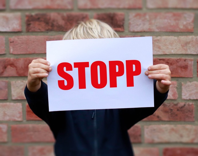Das Foto zeigt ein Kind, das einen Zettel vor sein Gesicht hält. Das Gesicht ist dadurch verdeckt. Auf dem Zettel steht in großen Buchstaben "Stop!"