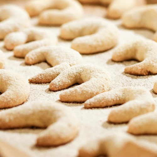 Das Foto zeigt ein Backblech, auf dem frisch gebackene und mit Puderzucker verfeinere Vanillekipferl liegen.